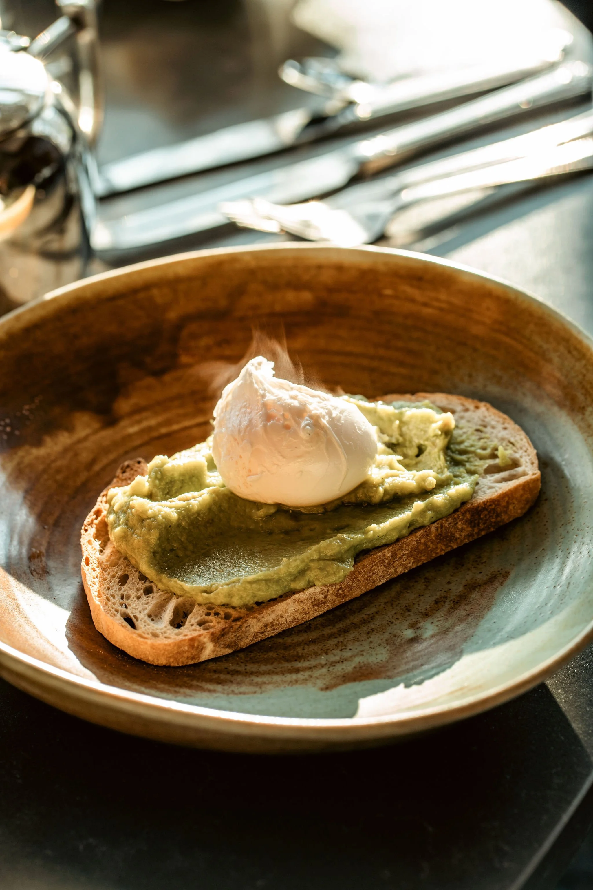 A slice of bread topped with mashed avocado and a poached egg on a brown ceramic plate.