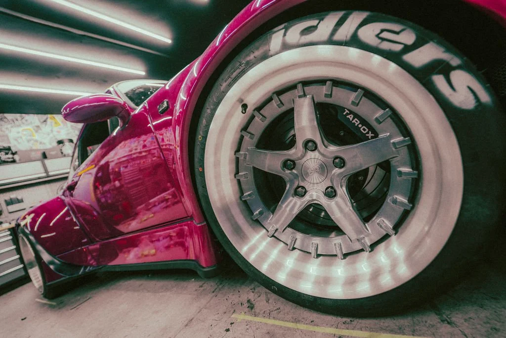 Close-up of a pink Porsche RWB with a low stance showing a large wheel with a unique rim design and a low-profile tire in a workshop environment.