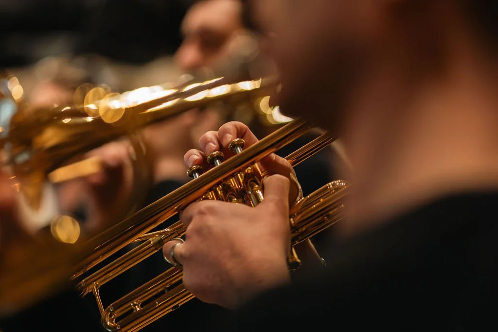 Close-up of a musician's hand playing a gold-colored trumpet during an orchestra performance, with other musicians blurred in the background.