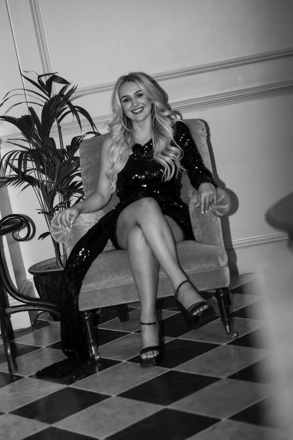 A young woman with long, wavy blonde hair, wearing a black sequined dress and high heels, sits on an upholstered armchair in a room with a checkered floor and decorative wall molding, smiling at the camera.