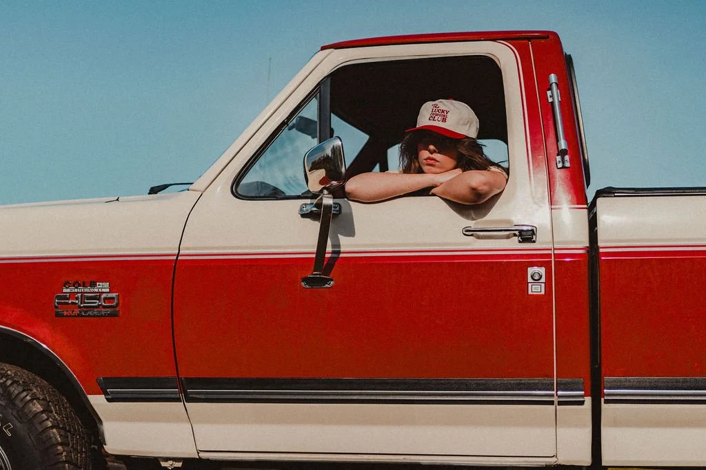 A woman with wavy brown hair wearing sunglasses and a gray cap with red writing, leaning on the window of a vintage red and cream Dodge Ram 150 pickup truck under a clear blue sky.