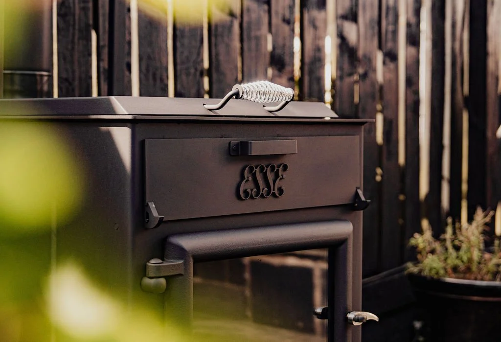 Black outdoor cast iron stove with a handle and decorative cutout on the door, set against a wooden fence, with some greenery in the foreground and a plant in a pot to the right.