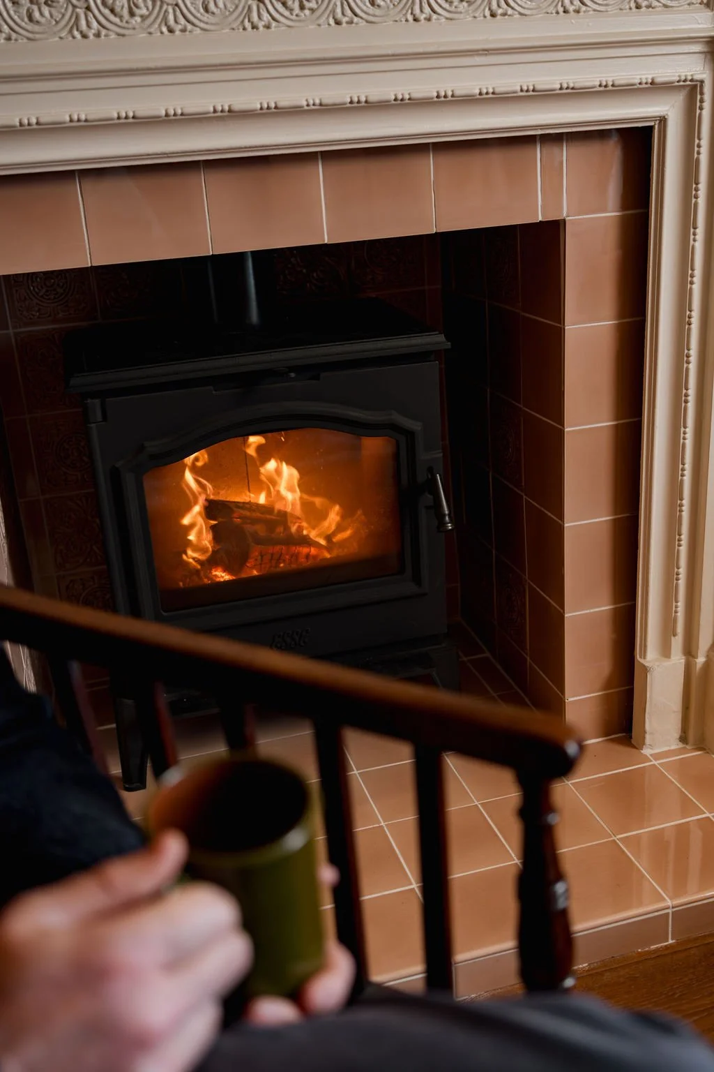 A cozy indoor scene with a person holding a mug near a fireplace with a burning fire, brown tiles around the fireplace, and decorative molding above.