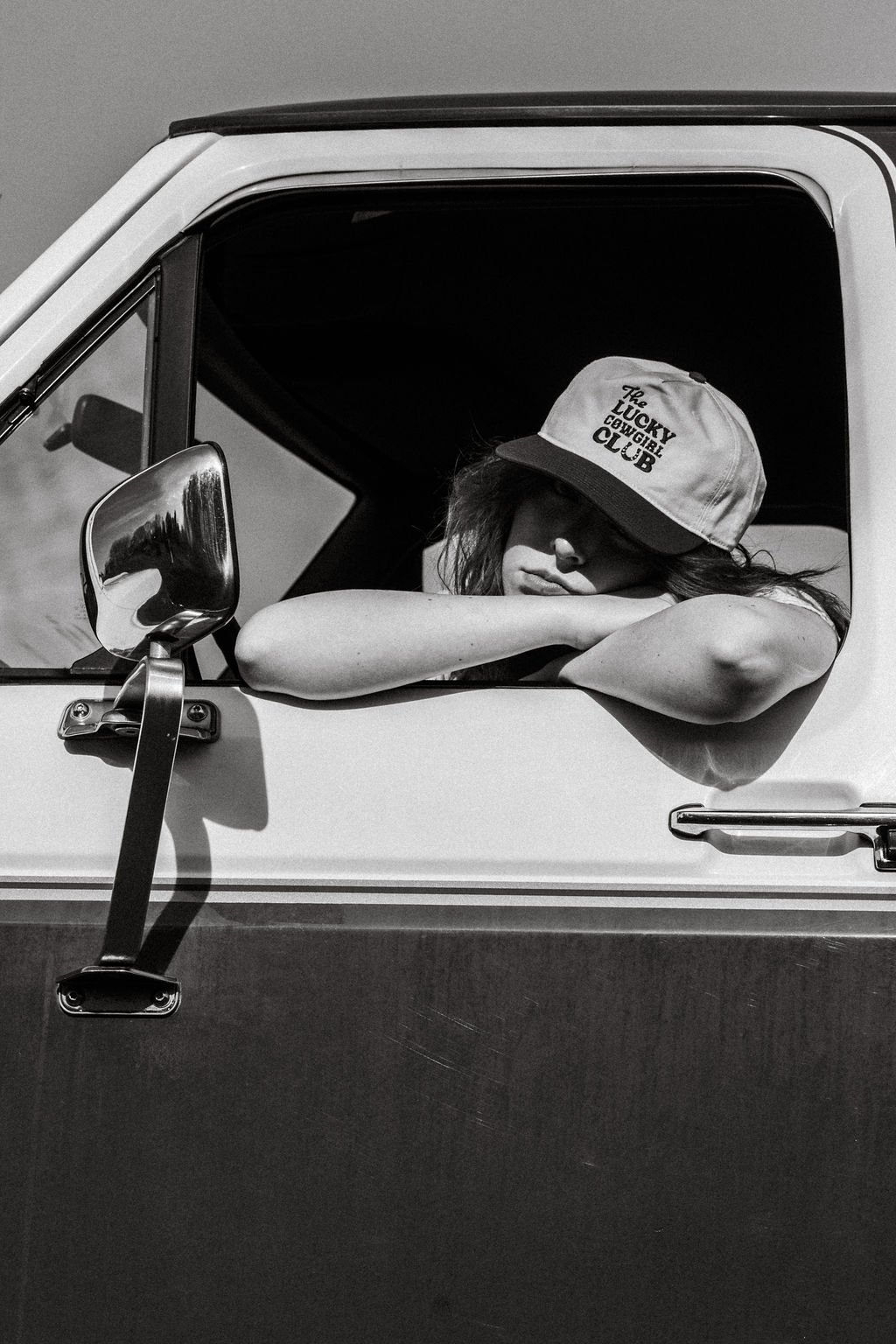 A young woman sitting in the driver’s seat of a vehicle, resting her arm on the window ledge, wearing a baseball cap that reads 'The Lucky Cowgirl Club', with her head tilted and eyes closed.