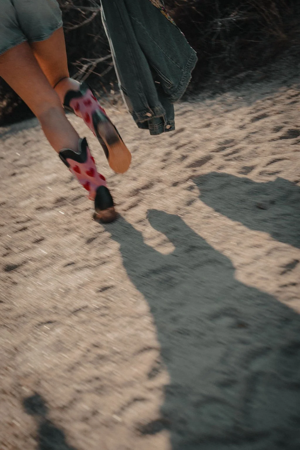 Close-up of a person's legs walking on sandy terrain, with a denim jacket hanging from their hand. The person's footwear has a ladybug pattern. Shadows are cast on the ground.