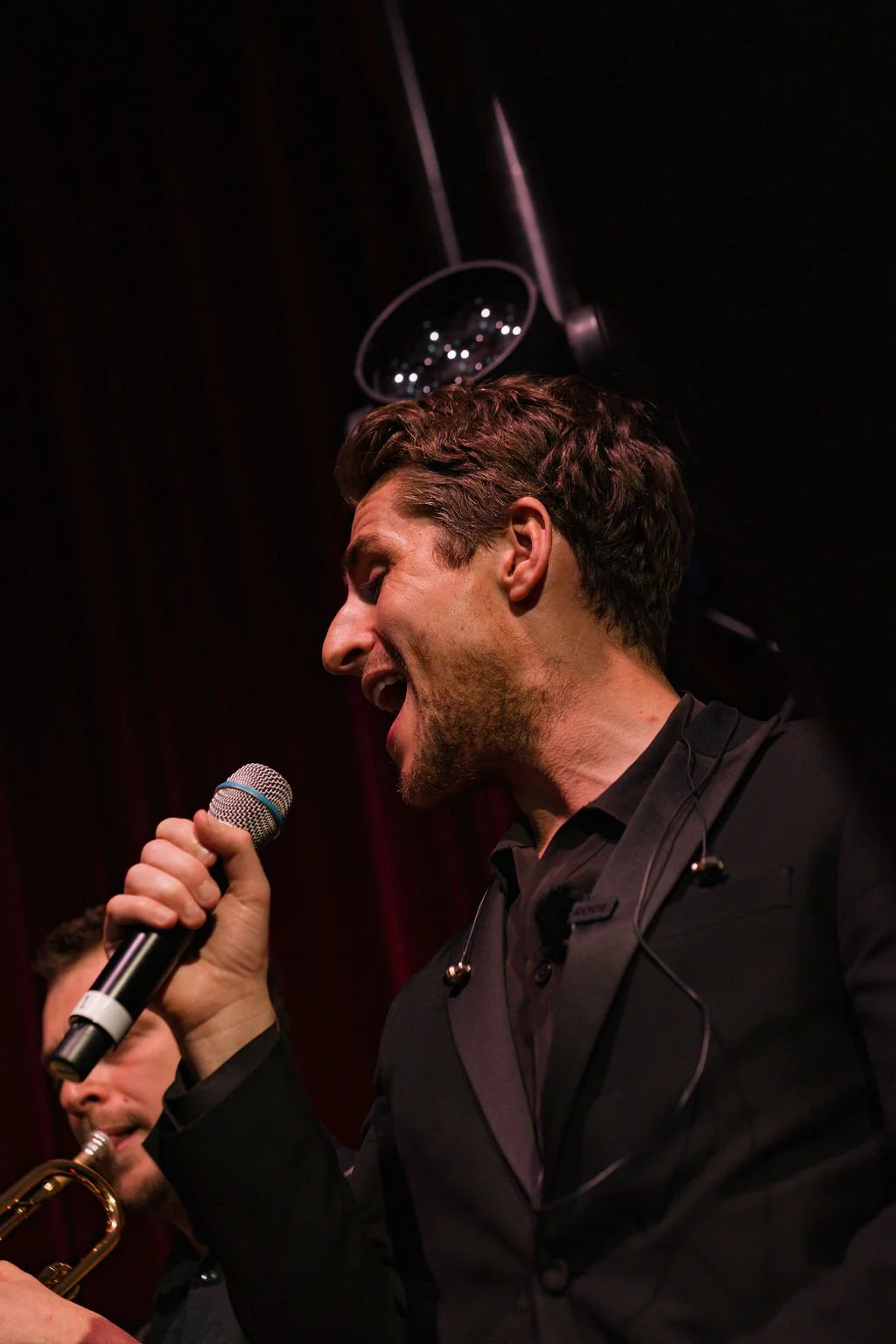 A man with brown hair singing into a microphone on stage with a musician playing a trumpet in the background.