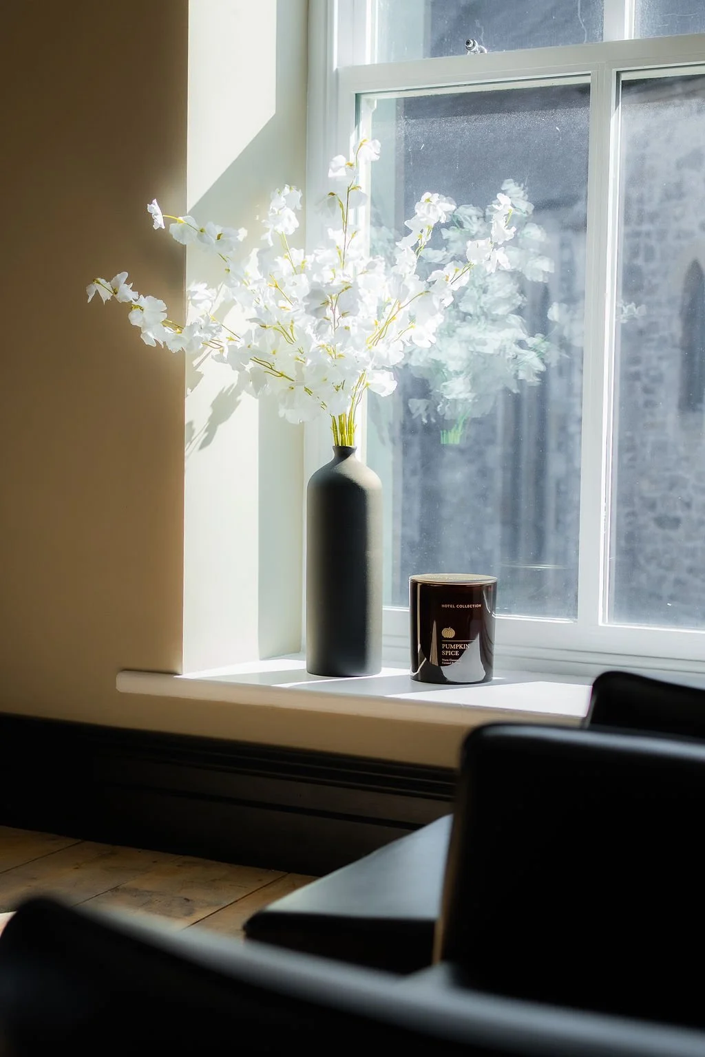 A black vase with white flowers on a windowsill, sunlight streaming in through a large window, with a black candle labeled "Pumpkin Spice" nearby.