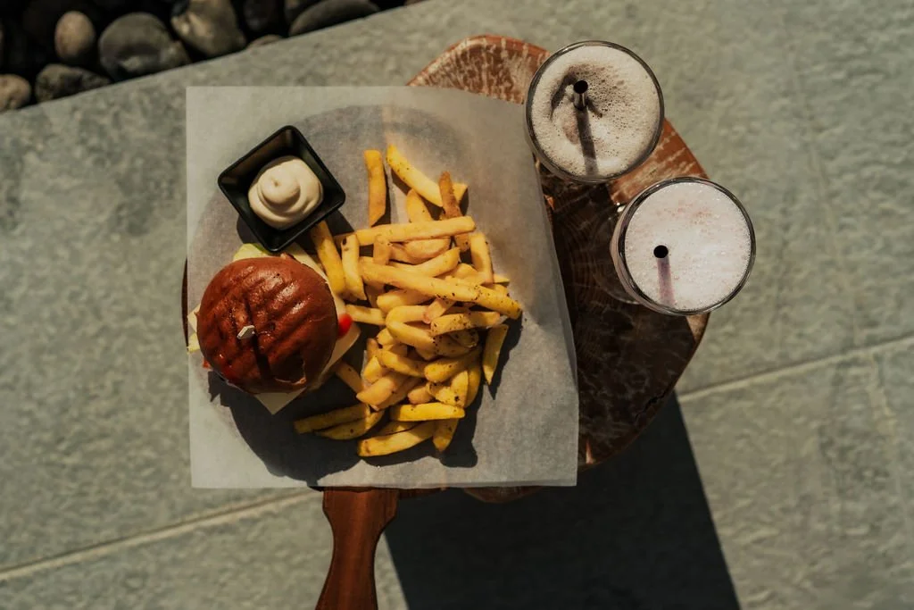 A top-down view of a meal with a burger, French fries with seasoning, a small container of mayonnaise, and two milkshakes on a small wooden table.