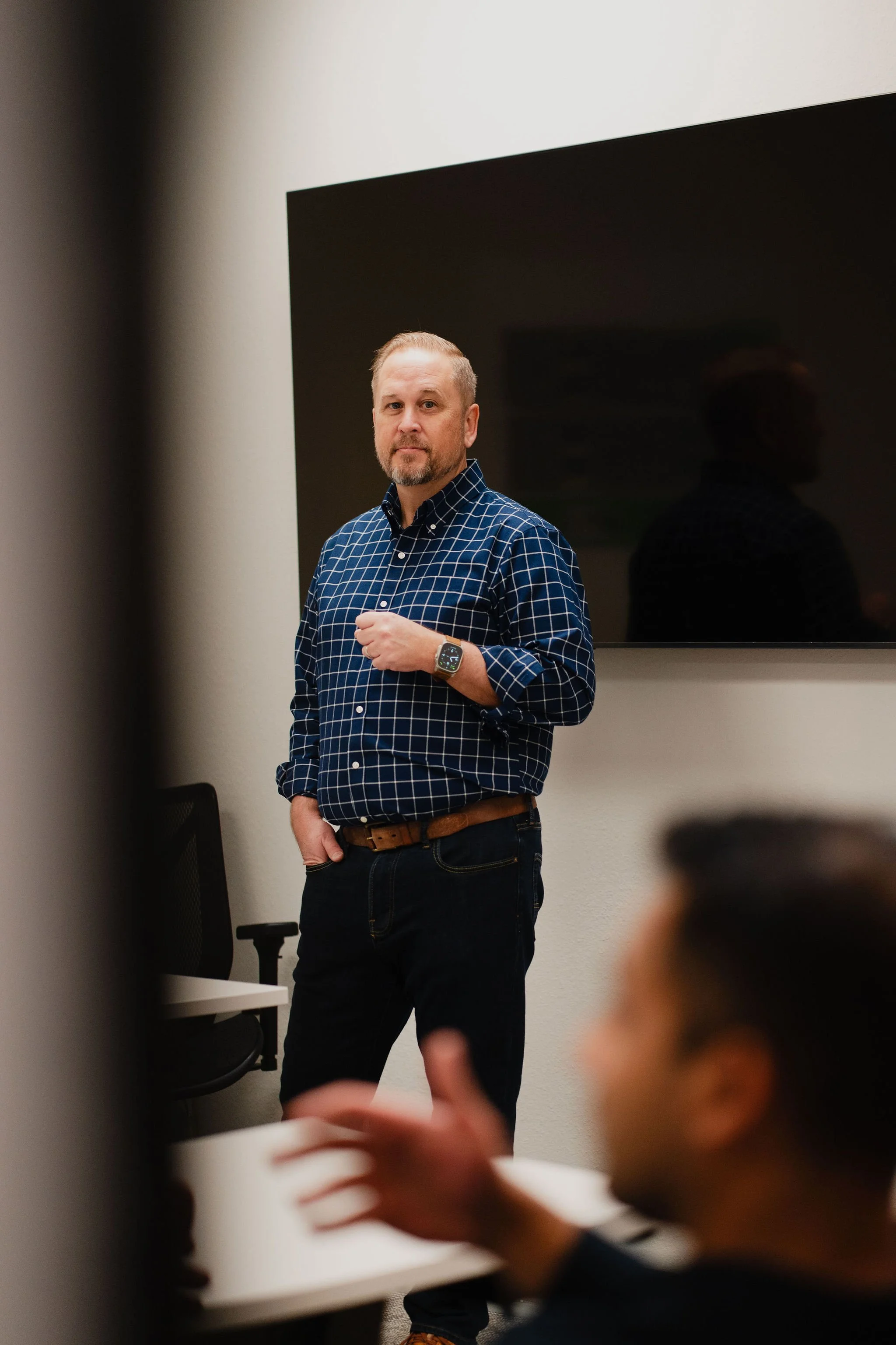 A man in a blue checkered shirt standing in front of a large flat screen television, with a blurred person in the foreground.
