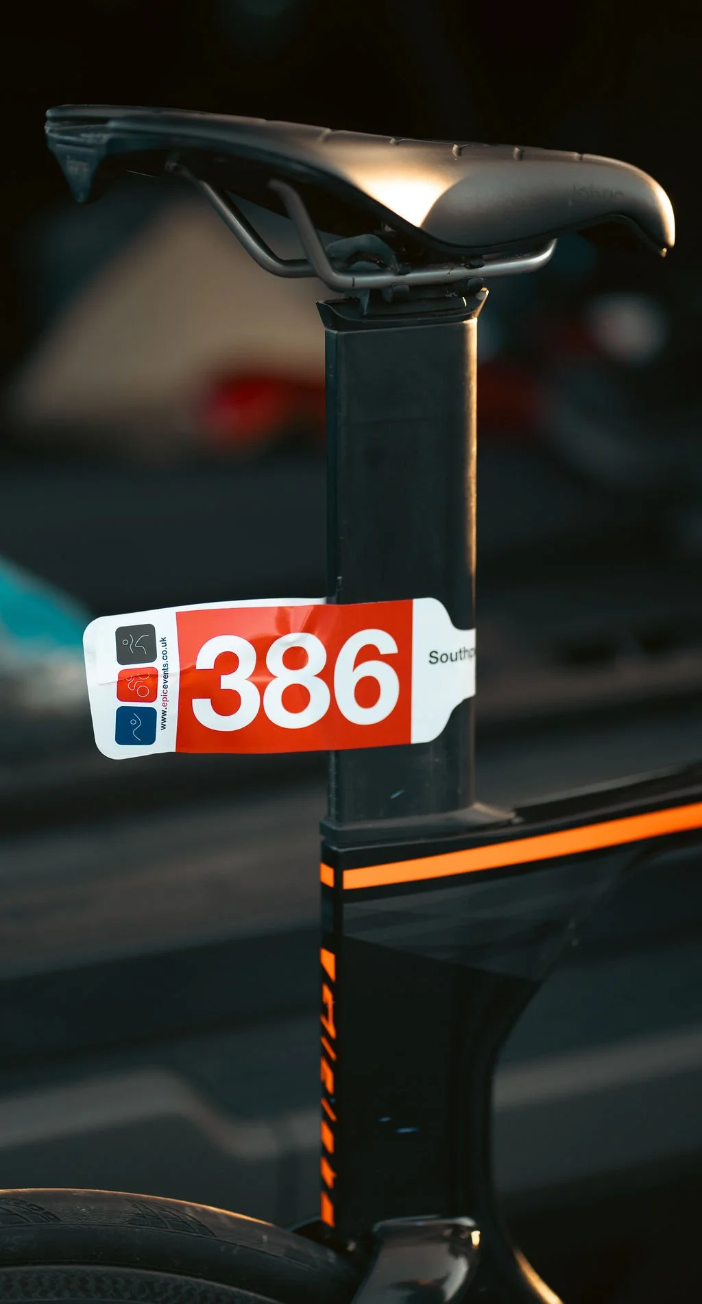 A bicycle saddle mounted on a seat post with a red and white number plate labeled '386' attached to the seat post. The background is blurred.