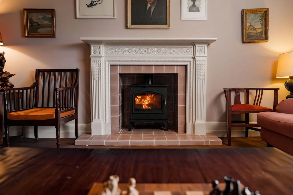 Living room with a decorative white fireplace mantel, a wood stove with fire, and framed artwork on the wall. Two chairs are positioned on either side of the fireplace, and a lamp illuminates the room.