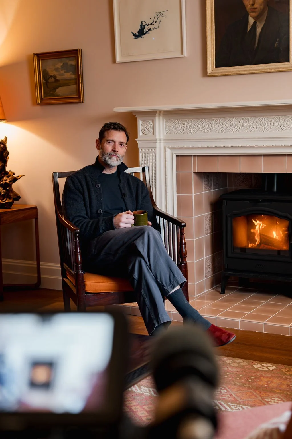 A man with a beard, sitting in a wooden chair with orange cushion, holding a mug, next to a lit fireplace in a cozy living room with framed artwork on the wall.