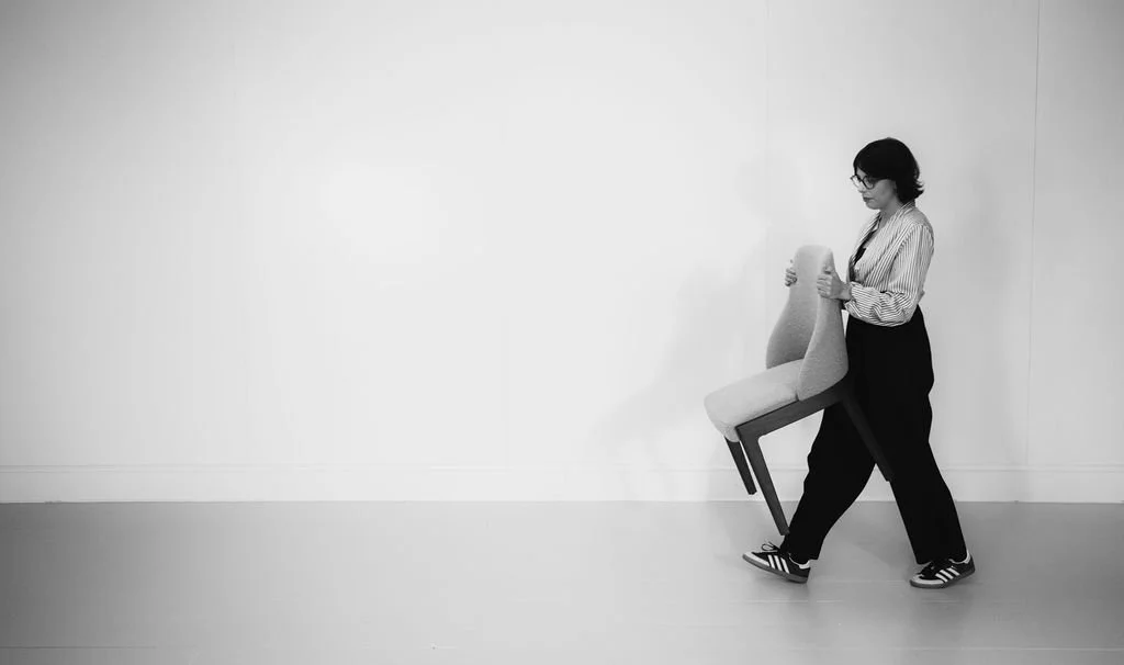 A woman with glasses and dark hair, wearing a striped shirt and black pants, is carrying a chair against a plain white wall.