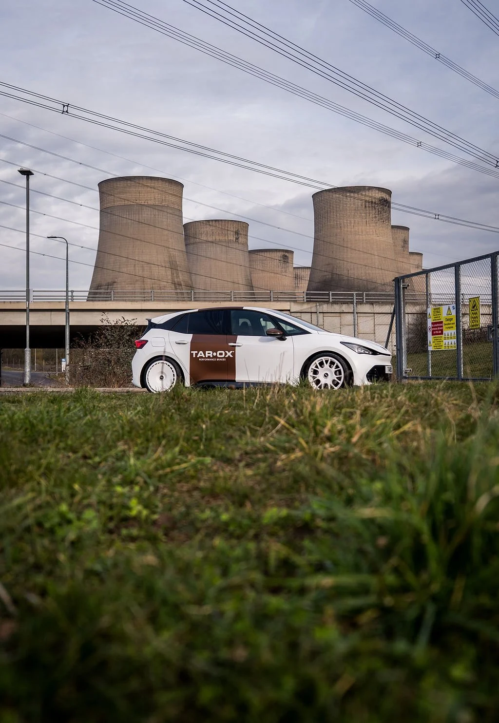 A white car parked on grass in front of a fence with warning signs, nuclear power plant cooling towers in the background, and power lines overhead.