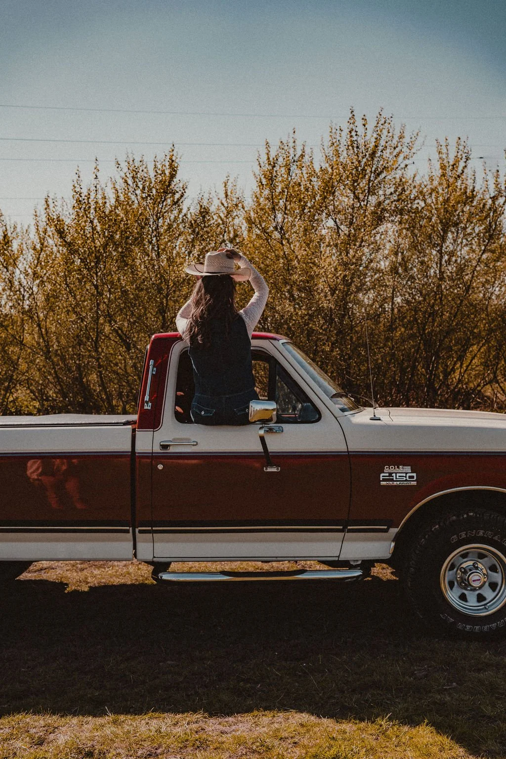 A woman sitting in the truck's cab with her back to the camera, wearing a wide-brimmed hat and looking at the sky, in a rural outdoor setting with autumn trees in the background.