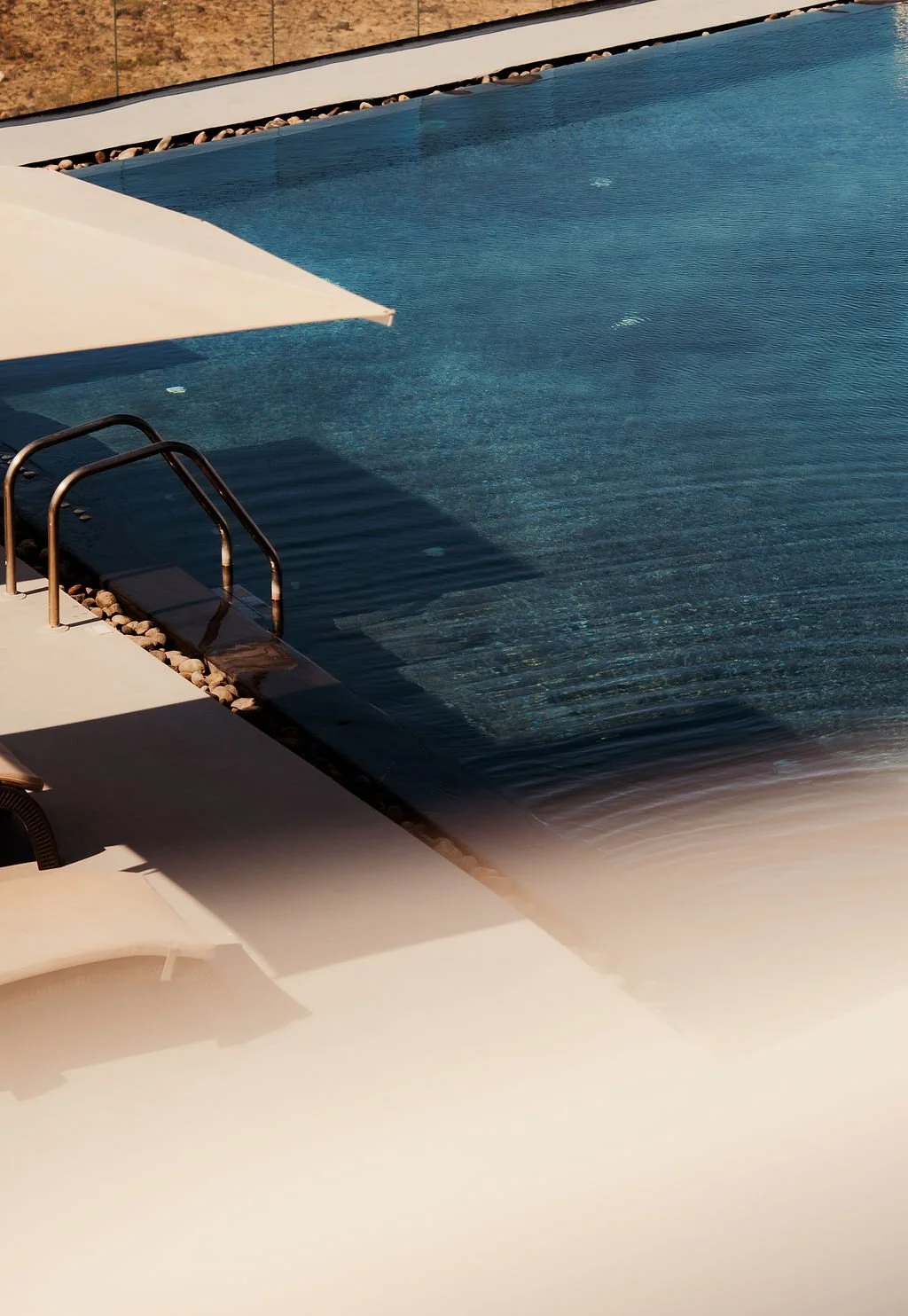 A swimming pool with metal handrails, a white poolside lounge chair, and a large umbrella. The pool is surrounded by rocks and a brown stone wall.