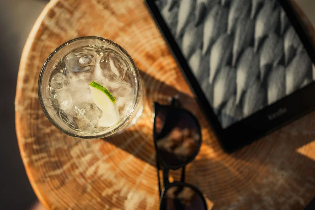 A glass of water with ice and a lime wedge on a wooden table, alongside sunglasses and a patterned box.