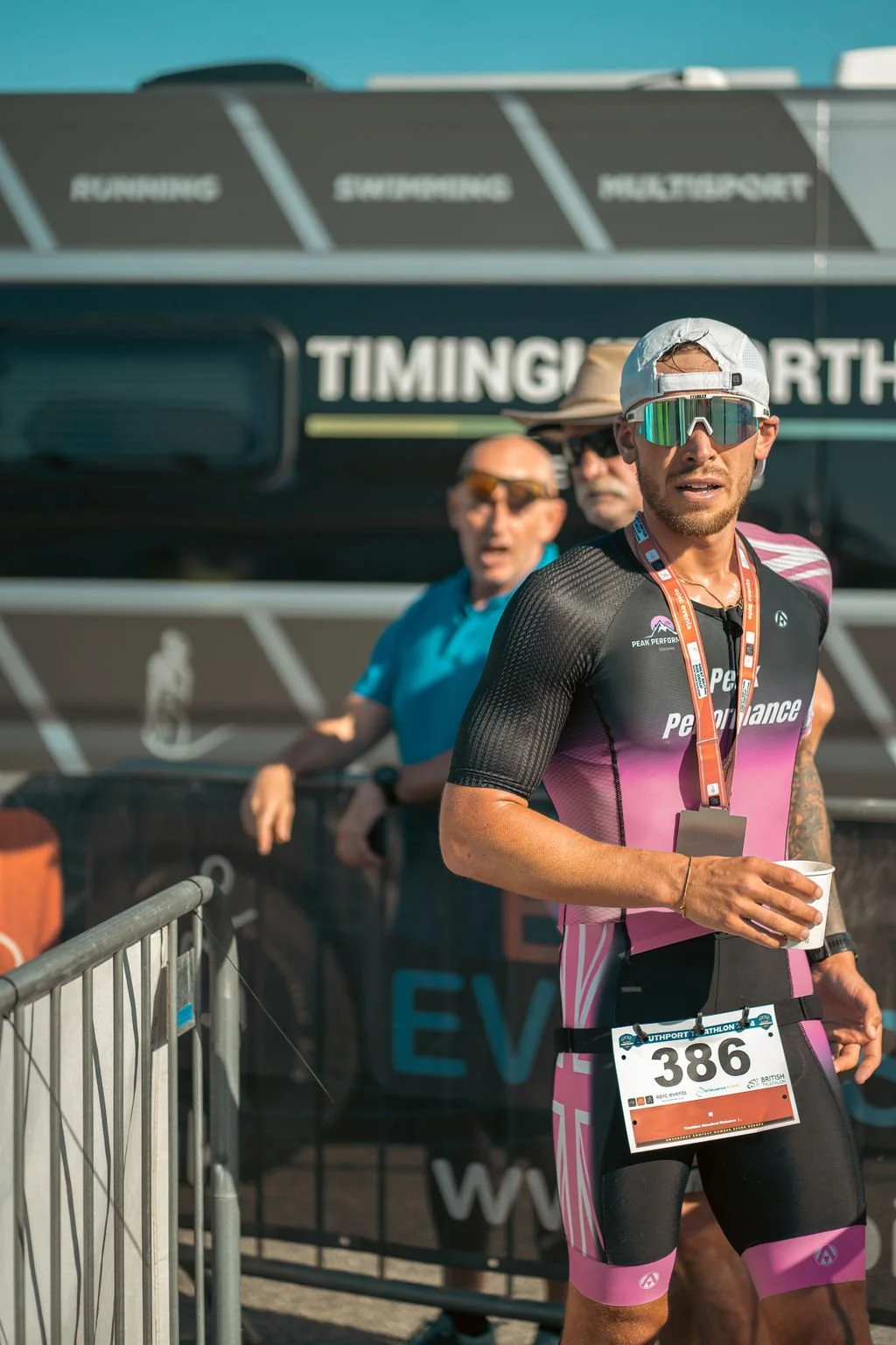 A male athlete in a pink and black performance suit with race number 386 wears sunglasses and a white cap, holding a cup at a race event, with a timing and sports support truck in the background.