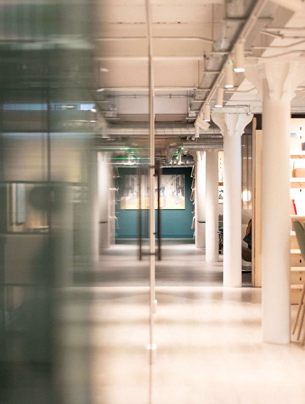 Empty modern office corridor with white columns, ceiling pipes, and art on the wall, viewed through a glass reflection.