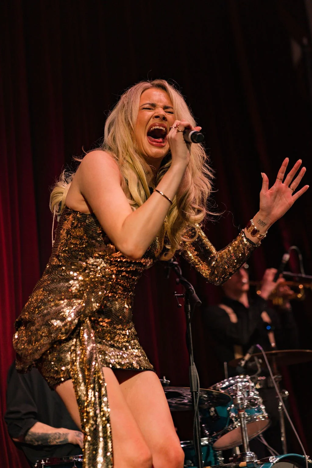 A female singer with long blonde hair performing on stage in a shiny, gold sequined dress, singing passionately into a microphone, with a red curtain in the background and a band playing behind her.