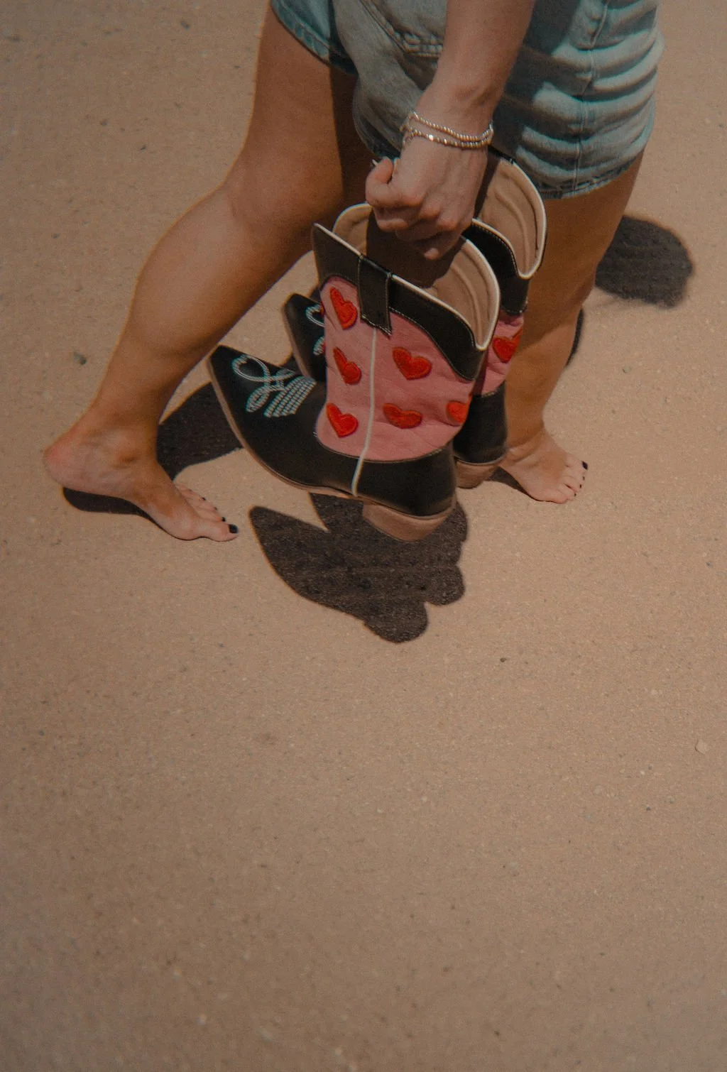 Person holding a small handbag with red heart designs, walking barefoot on sandy ground, wearing denim shorts and bracelets.