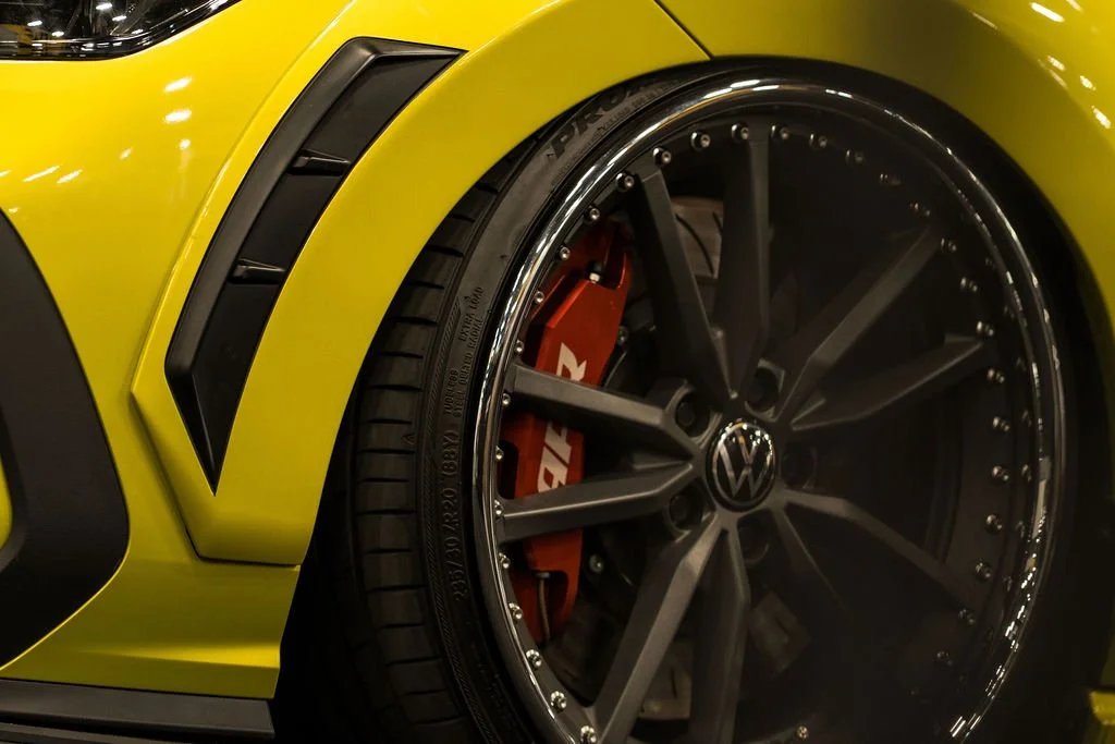 Close-up of a yellow sports car's front wheel, featuring a black multi-spoke rim, a red brake caliper with white lettering, and part of the car's yellow body with black accents.