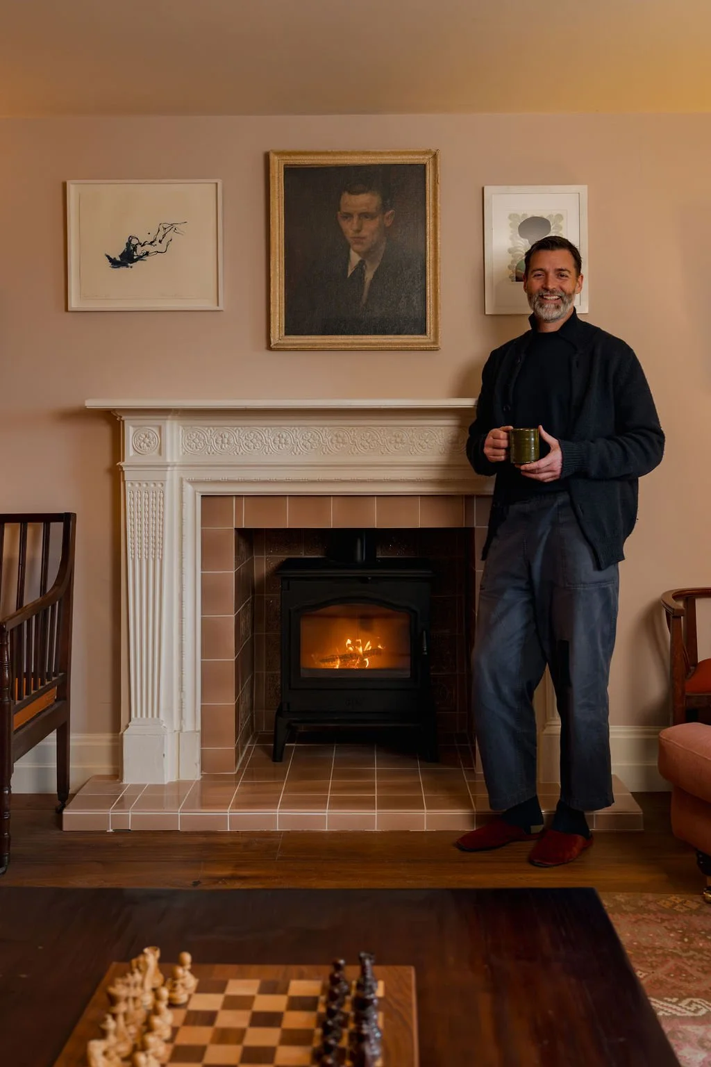A man with a beard standing in a living room, holding a mug, smiling, near a fireplace with a fire burning. The wall behind him has three framed artworks, including a portrait of a young man, abstract and line art pieces.