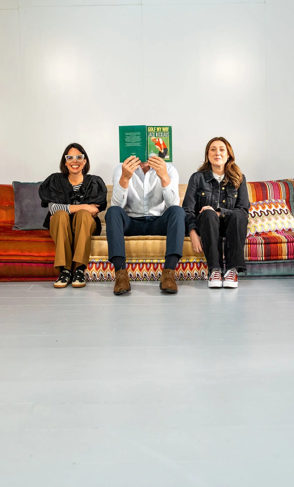 Three people sitting on a colorful couch against a white wall, one covering face with a book, all smiling and casually dressed.