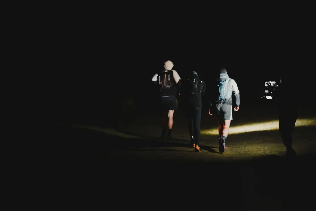 A group of four people hiking at night, illuminated by their headlamps, walking on a trail in a dark outdoor environment.