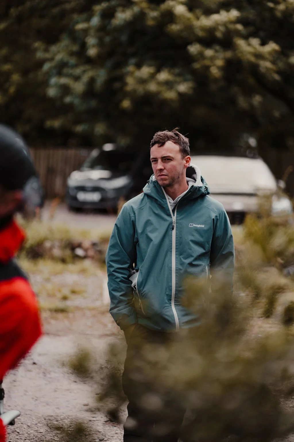 A man in a teal Berghaus waterproof jacket stands outdoors with his hands in his pockets, looking to his right. He appears serious, with short brown hair and slight facial stubble. Behind him, there are blurred vehicles and a large tree.