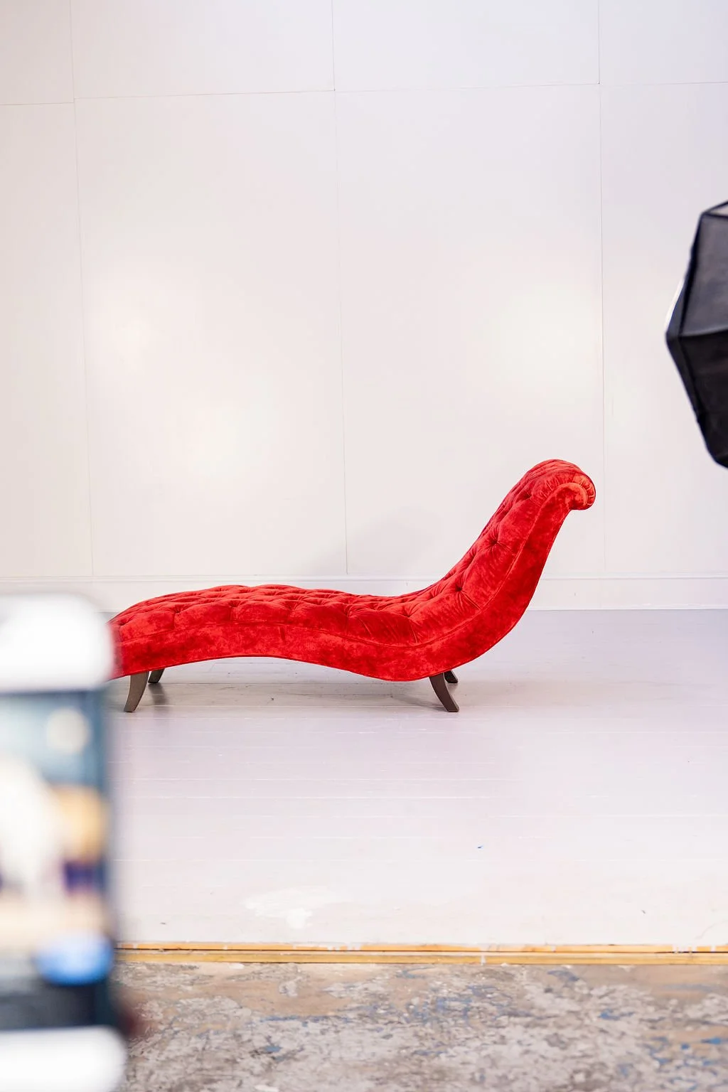 A red chaise lounge with a tufted design and curved backrest in front of a plain white wall.