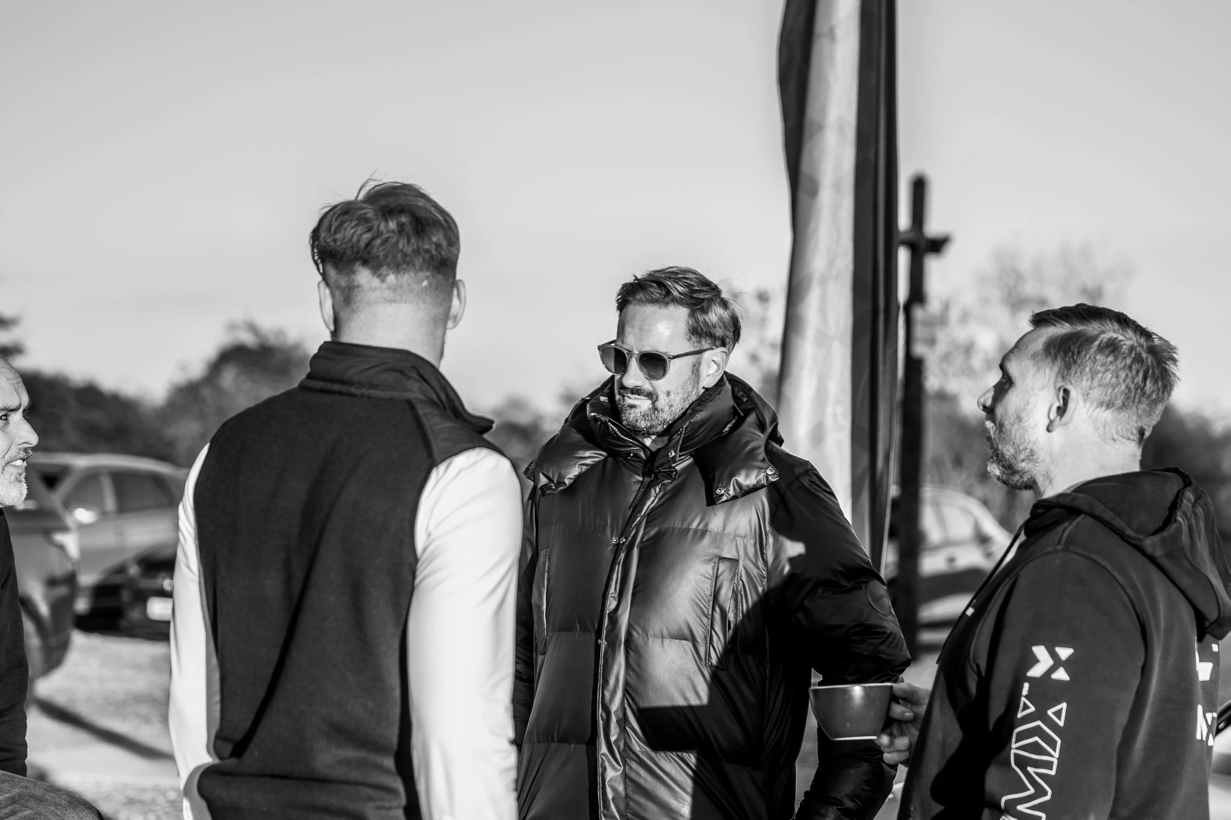 Four men having a conversation outdoors, one wearing sunglasses and a puffy jacket, others dressed casually, cars in the background.