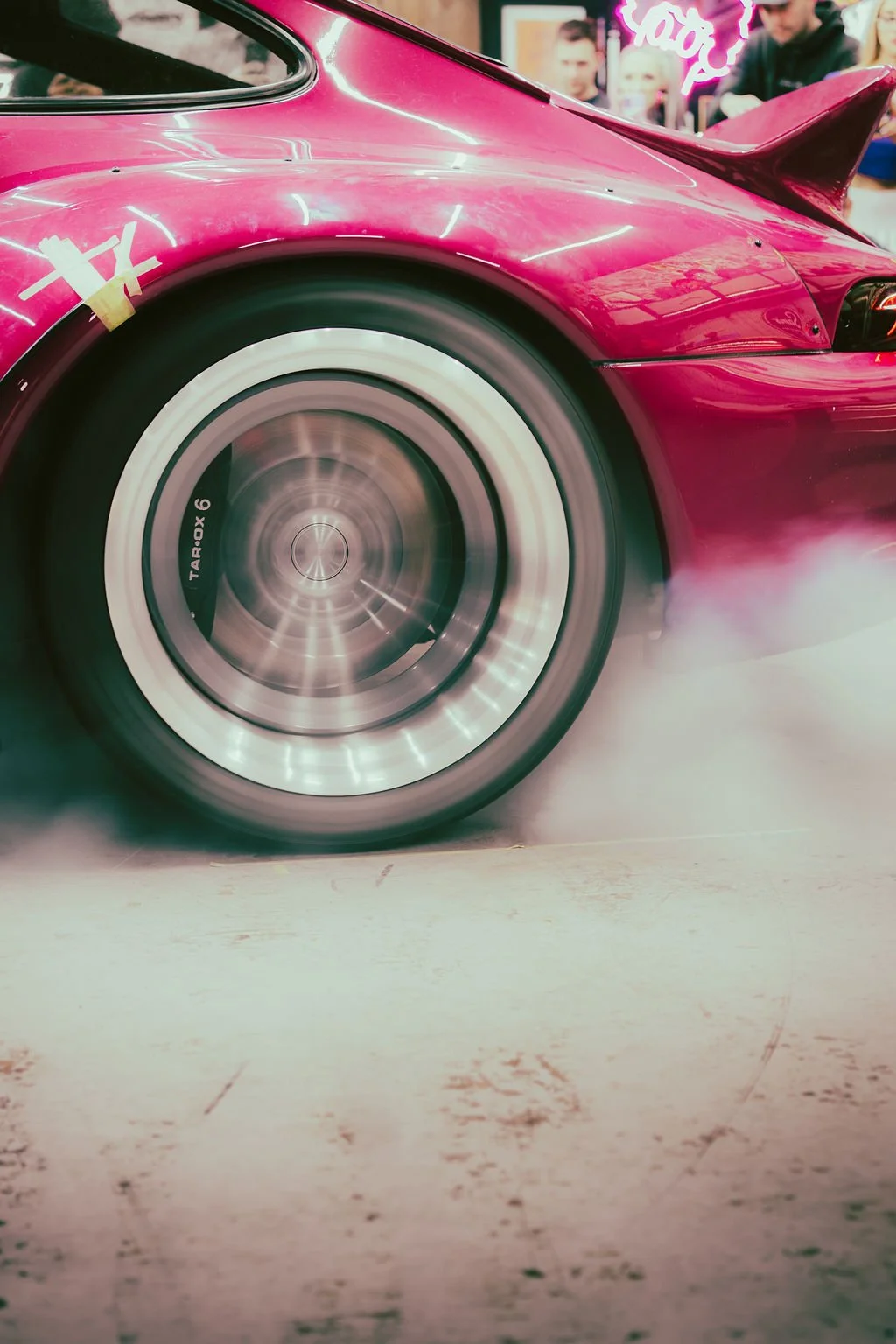 Close-up of a pink Porsche RWB car's tire spinning, with smoke coming from the rear tires, and a crowd of people in the background at an indoor event.