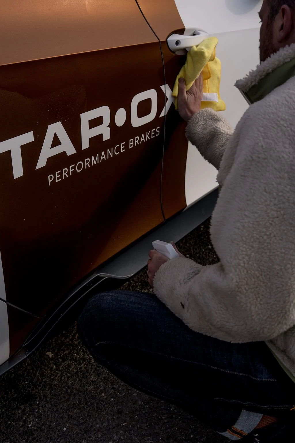 Person cleaning a car door with a yellow cloth, near a logo that reads 'TAR•O PERFORMANCE BRAKES'.