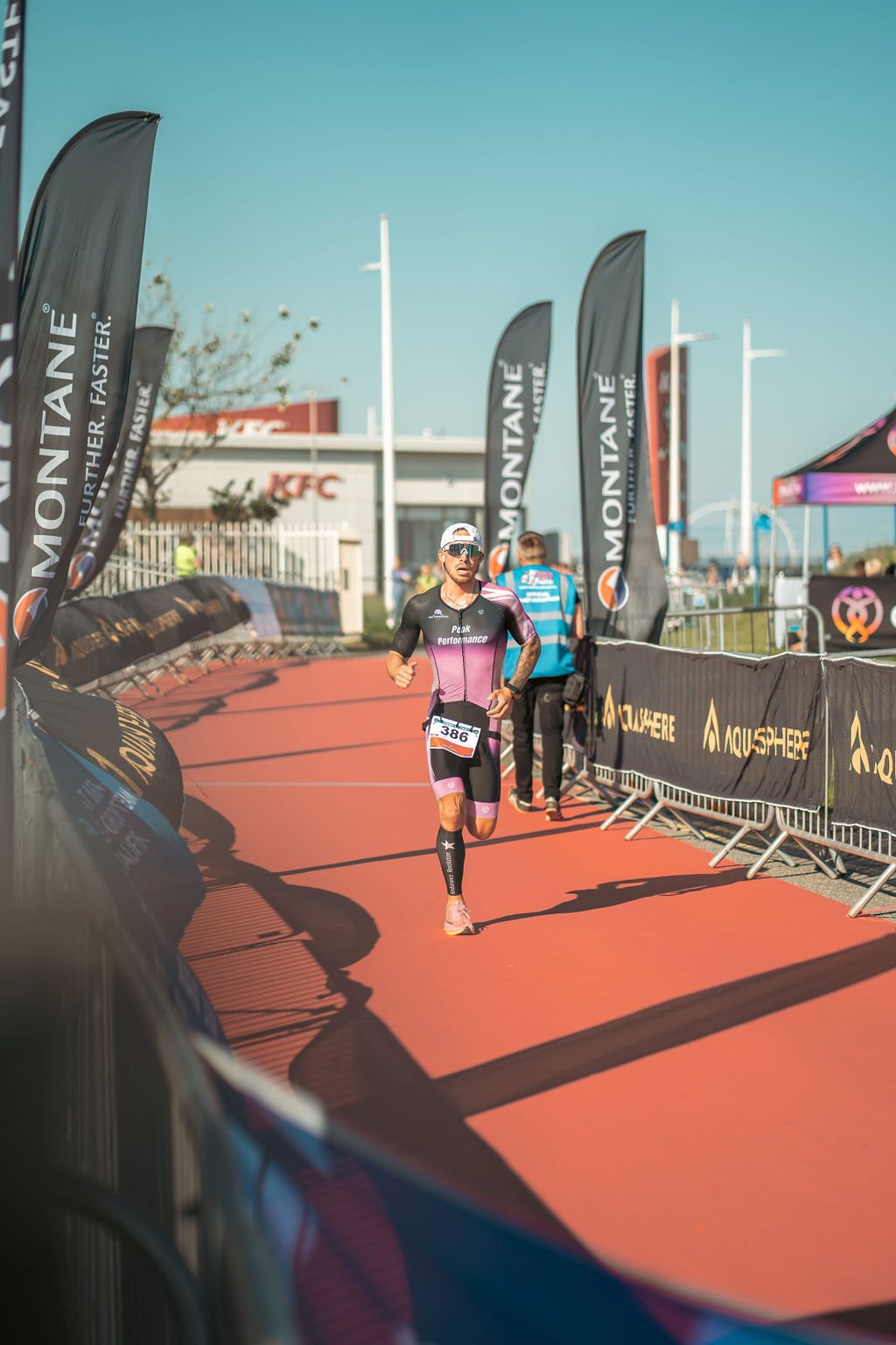 A male athlete running outdoors during a race, wearing a black and pink triathlon suit, sunglasses, and a race bib with the number 386. The race is taking place on a red carpet alongside sponsor banners and flags, with a building and a blue sky in th