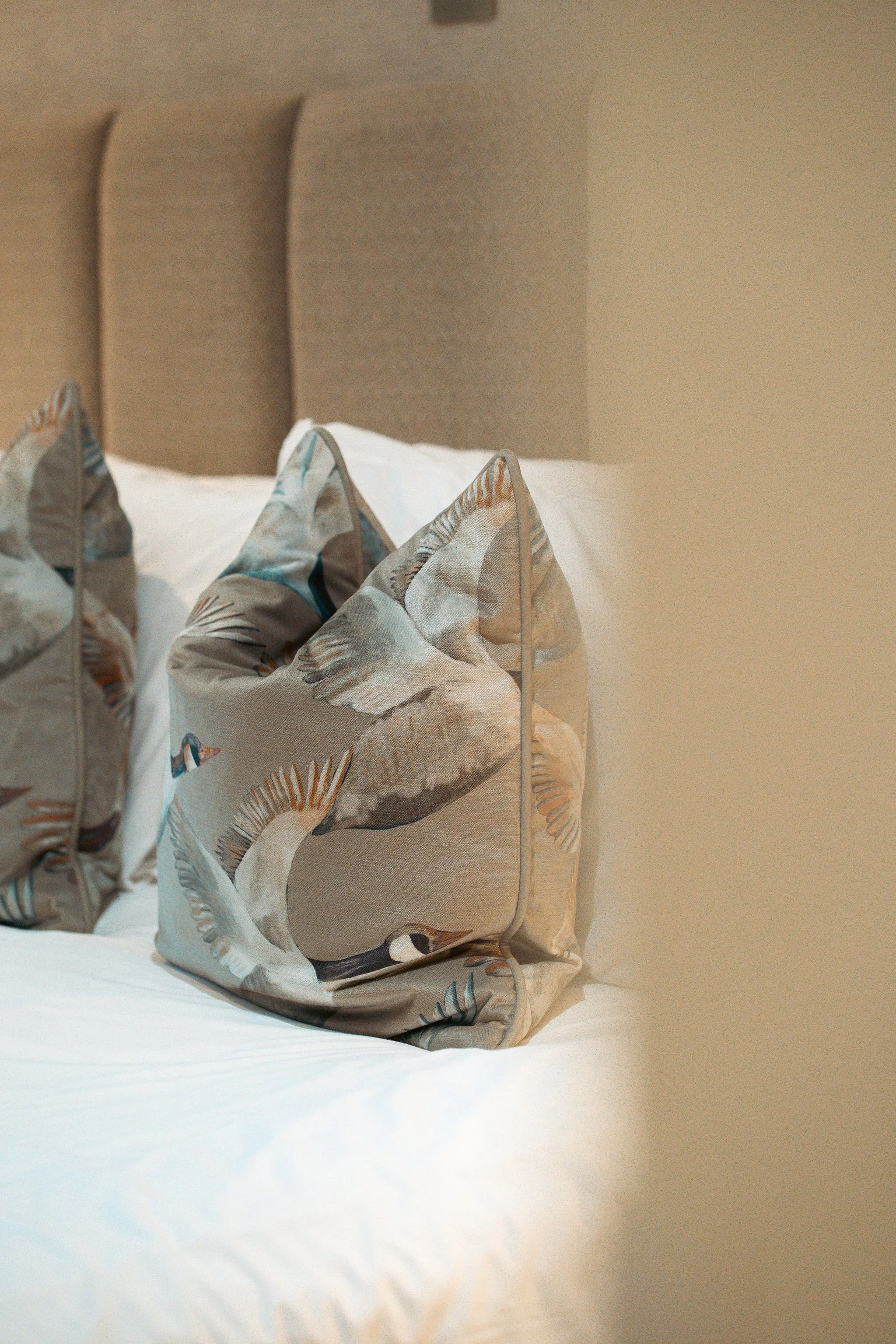 Decorative pillow with a bird and feather design on a bed with a beige headboard.