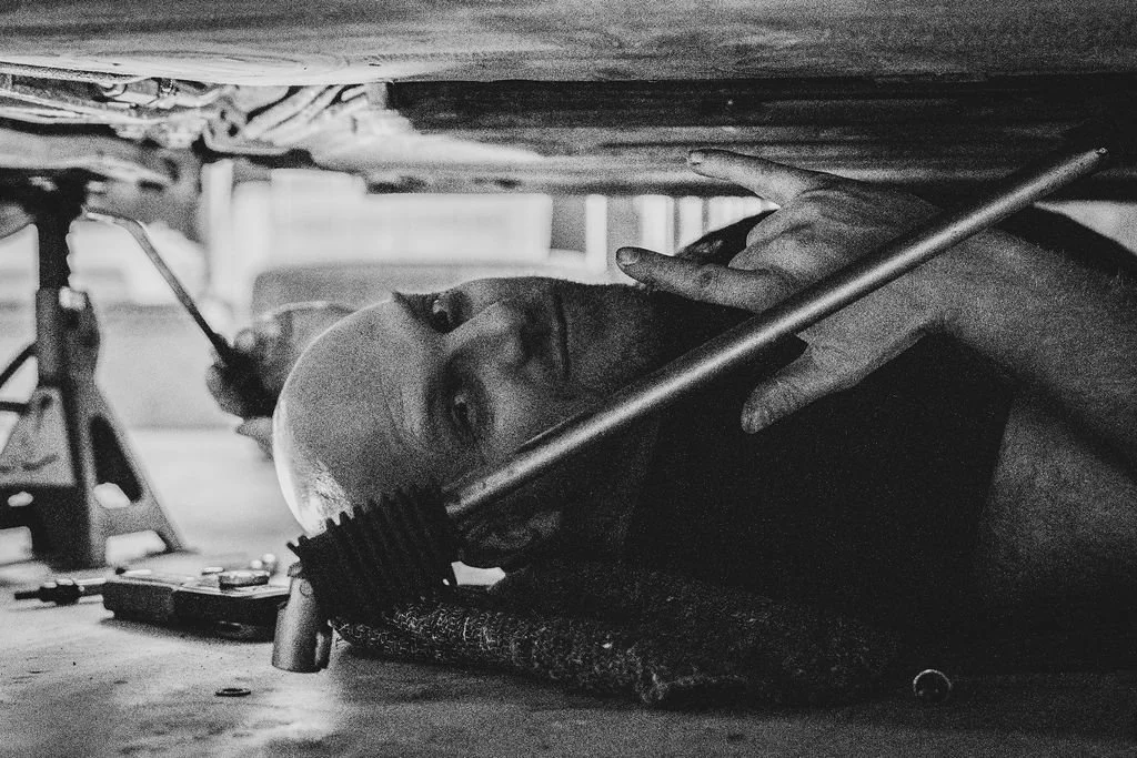 A person lying on the ground under a car working on something with tools, holding a long metal rod, with tools and equipment around.