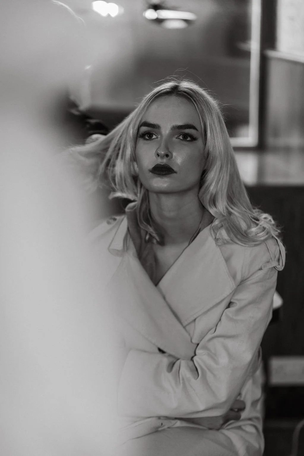 A black and white photo of a woman with blonde hair and dark lipstick, looking into the distance. She is wearing a trench coat and is seated indoors.
