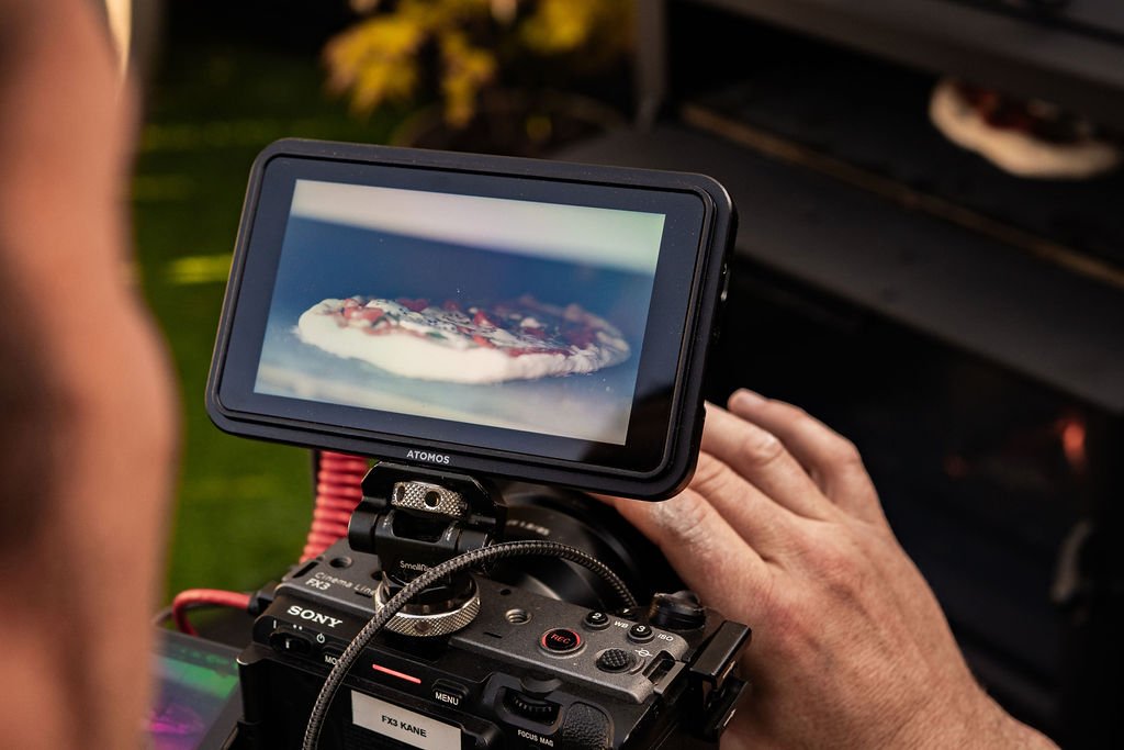 A person operating a Sony camera with a mounted external monitor, capturing a close-up of a pizza.