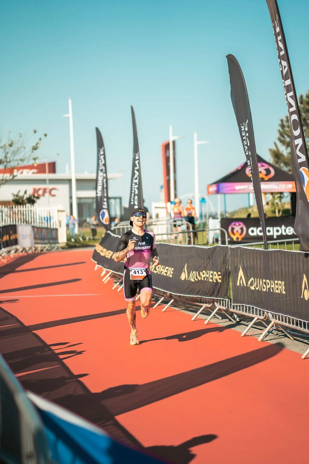 Female athlete running on a red race track, smiling, wearing black and pink athletic gear, sunglasses, and a race bib number 413, during a marathon or triathlon event.