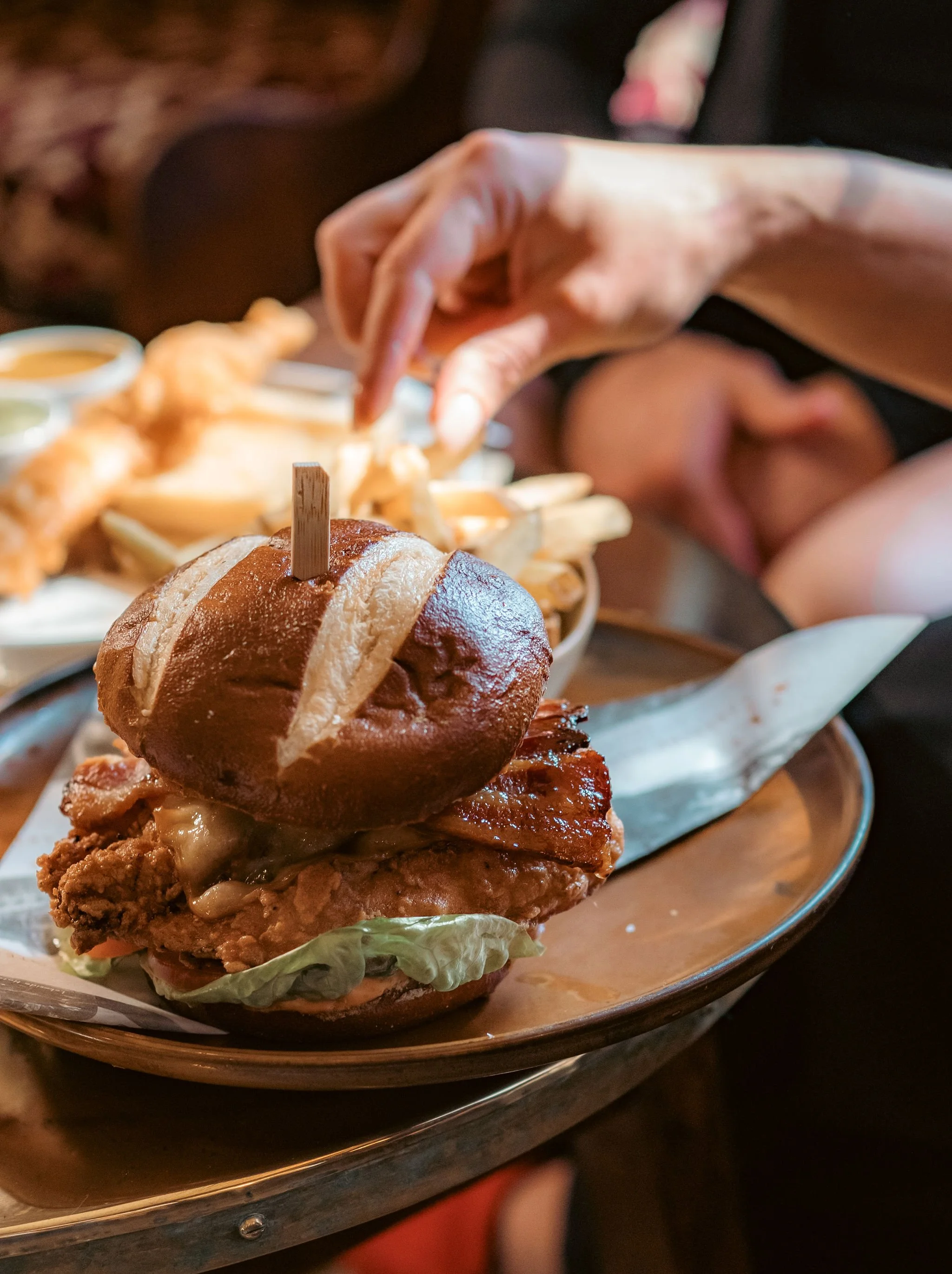 Close-up of a crispy fried chicken sandwich with lettuce, cheese, and a pretzel bun. In the background, a person is reaching for fries with dipping sauces.