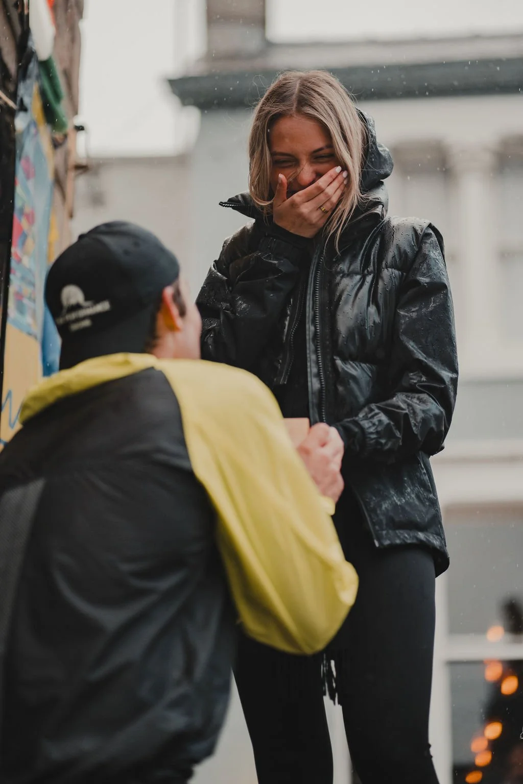 A woman in a black rain jacket is laughing and covering her mouth with her hand while talking to a person sitting down.