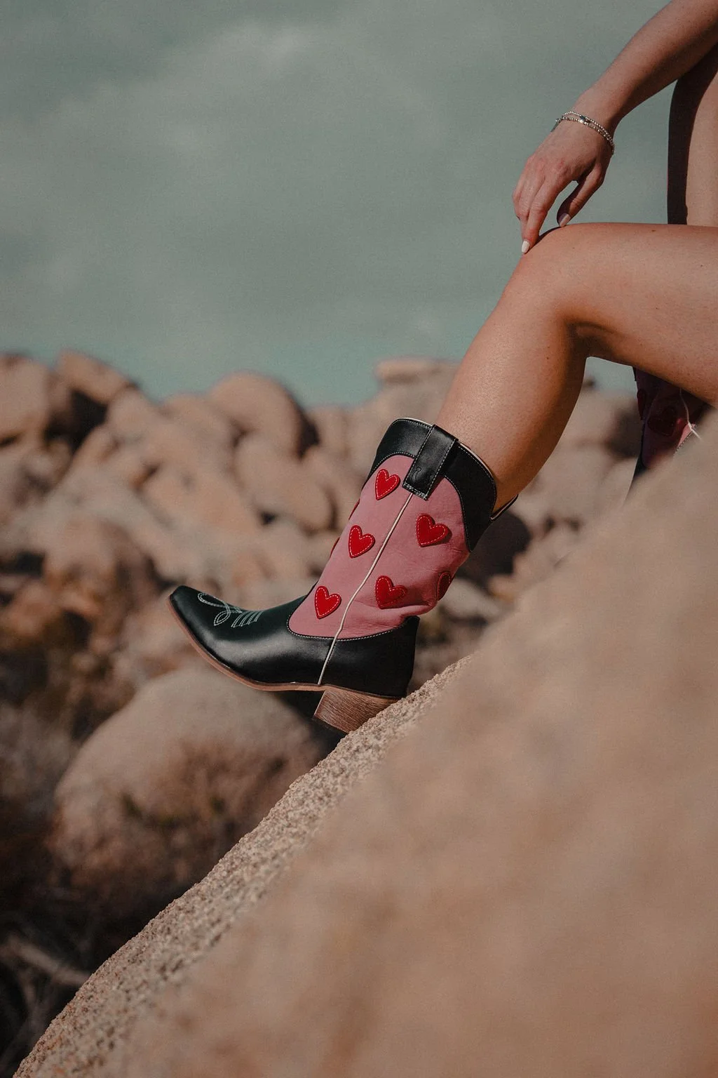 A woman wearing cowboy boots with pink and red heart designs, sitting on rocks near the beach, with her leg extended and hand resting on her knee.