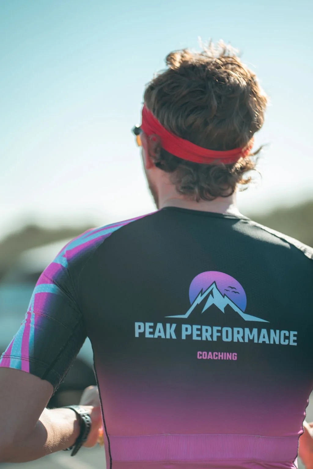 Back of a man wearing a black athletic shirt with a mountain and moon logo that reads 'Peak Performance Coaching' and a red headband, outdoors on a sunny day.