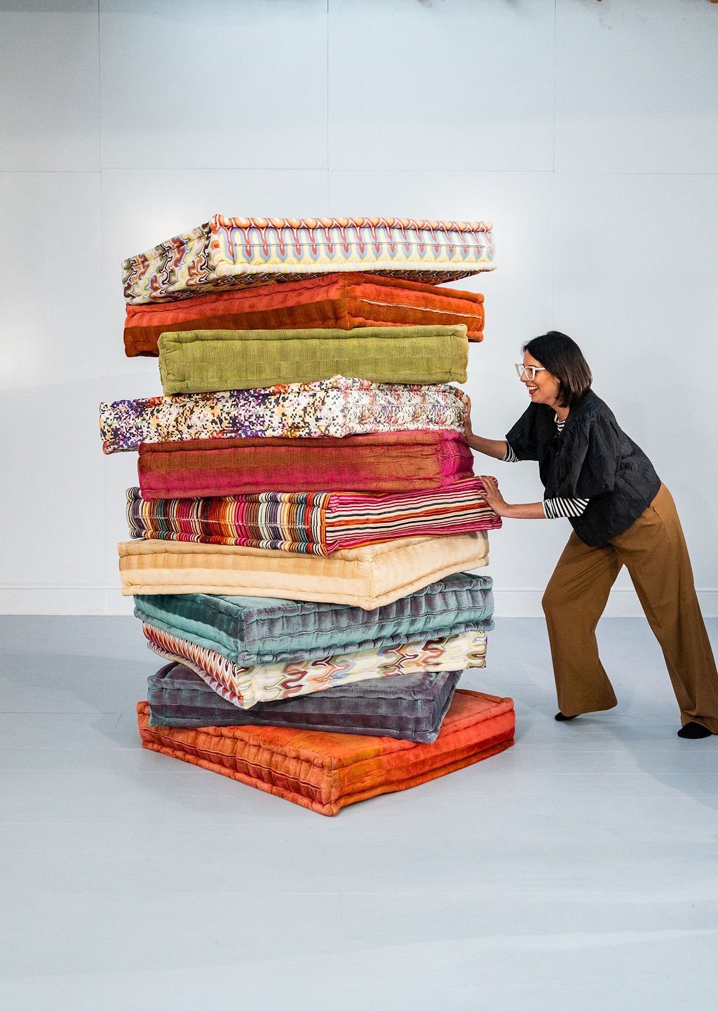 A woman wearing glasses and brown pants is pushing a large stack of various colorful, patterned cushions in a white-walled room.