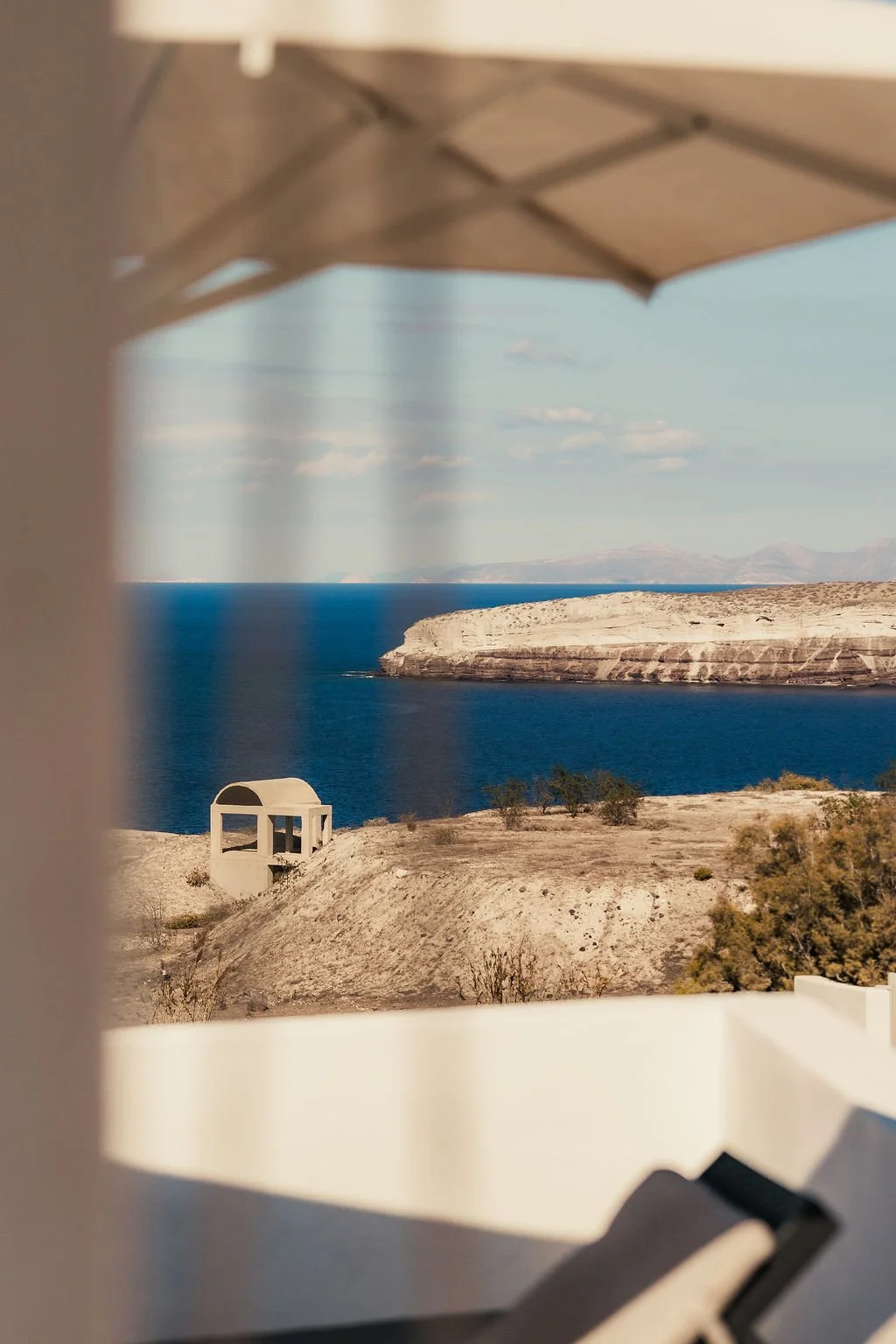 Scenic view of a large body of water, cliffs, and distant mountains seen through a window with a white frame and a white umbrella outside.