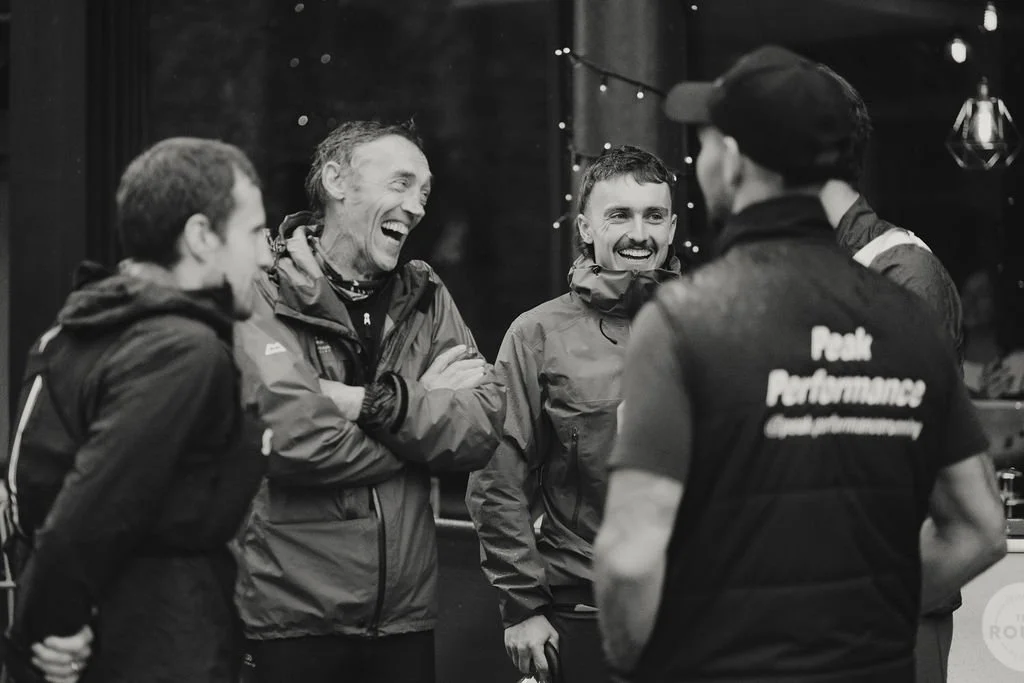 Four men in outdoor jackets are smiling and laughing together inside a dimly lit room with string lights, engaging in conversation.