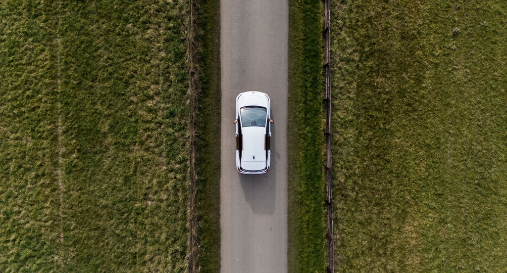 A white car driving down a narrow dirt road with grassy fields on both sides, viewed from above.