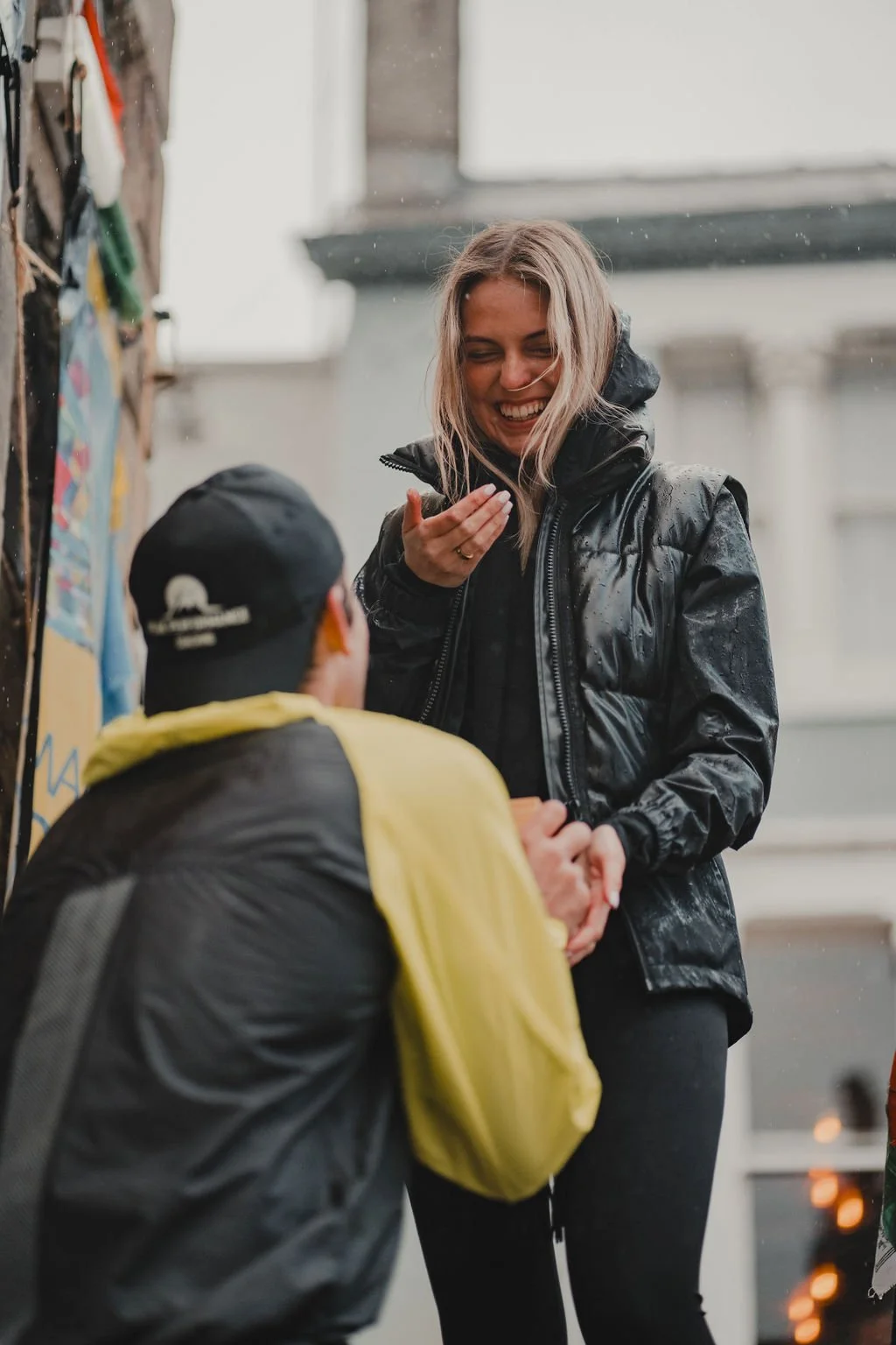 A woman laughing and holding hands with a man who's kneeling, outdoors in rainy weather, both dressed in black waterproof jackets, near a climbing wall.