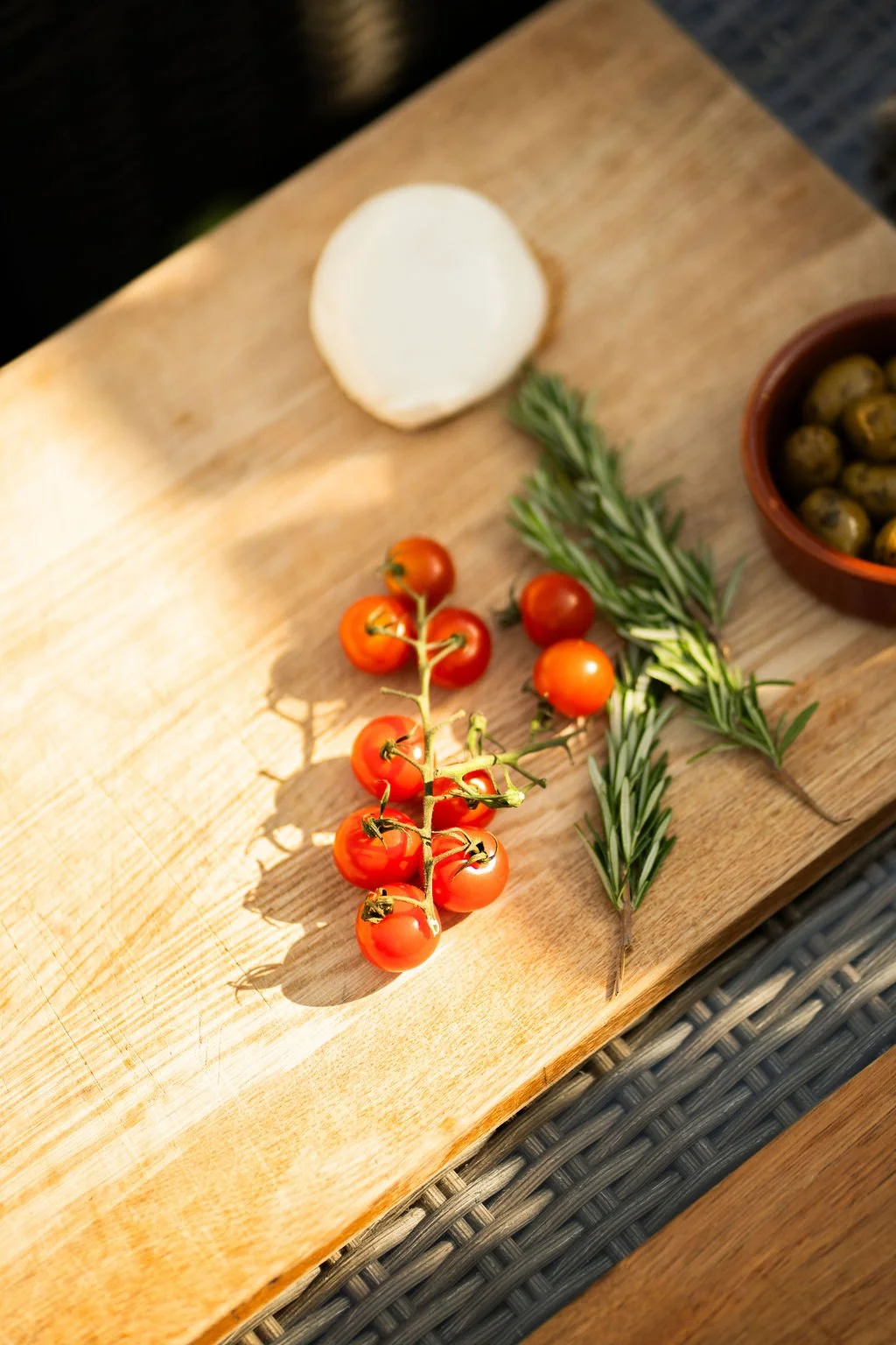 Cherry tomatoes, sprigs of rosemary, a slice of mozzarella cheese, and a bowl of green olives on a wooden cutting board.